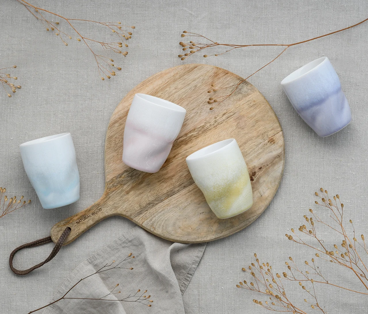 Vier creaTable Kaffeebecher »Squeezy Maytim« in Hellblau, Rosa, Gelbgrün und Lila, mit unregelmäßiger Form, stehen auf einem runden Holzbrett auf einem hellgrauen Leinentuch, umgeben von trockenen Ästen.