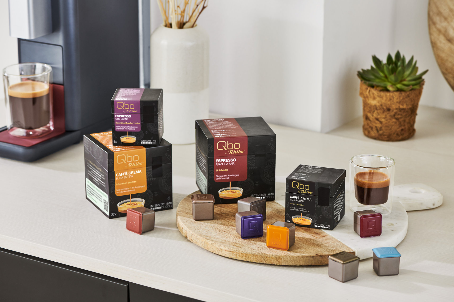 Auf einem Tisch stehen Qbo Espresso APANECA ANA Kapseln und eine Kaffeemaschine mit Espresso in einer Tasse.