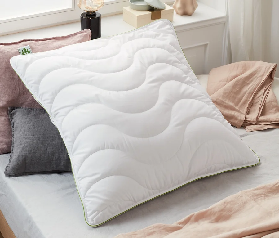 Auf einem Bett liegen ein weißes irisette® greenline Kopfkissen, ca. 80 x 80 cm, mit Steppung und grüner Bordüre, sowie graue und rosa Kissen.