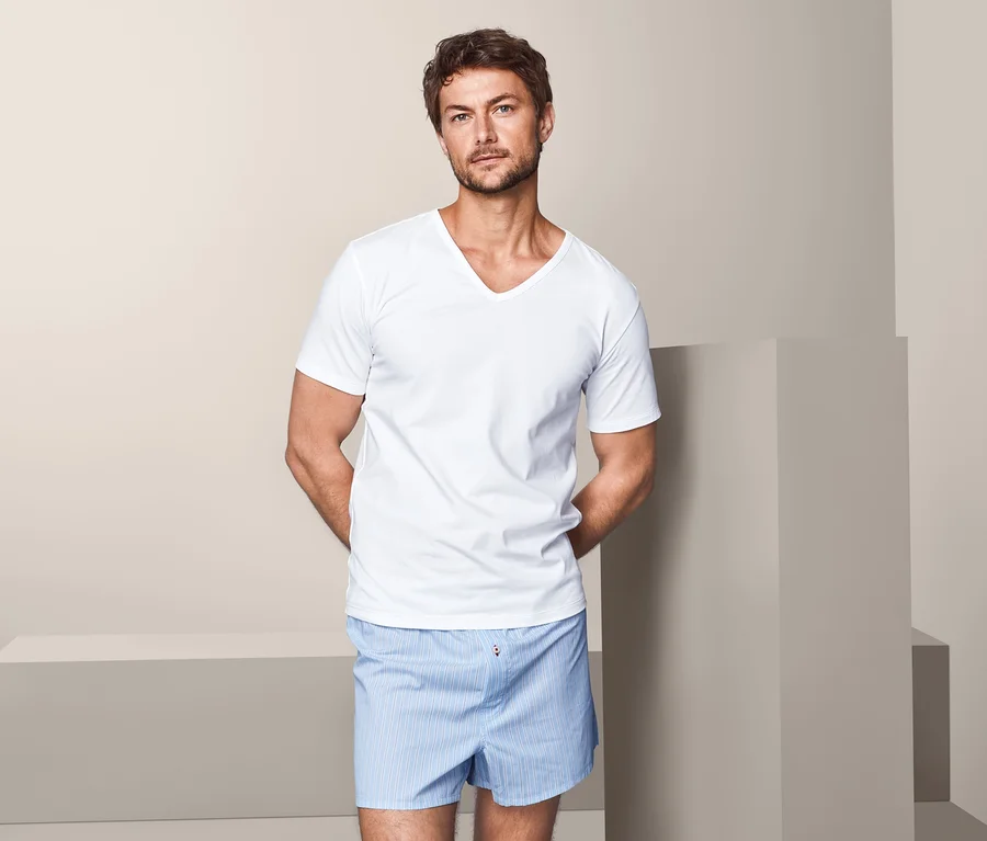Ein Mann posiert vor einem beige Hintergrund und trägt ein weißes Kurzarm-T-Shirt und blau-gestreifte Shorts. Das T-Shirt ist aus einem 2er-Pack Jersey-Halbarm-Unterhemden.