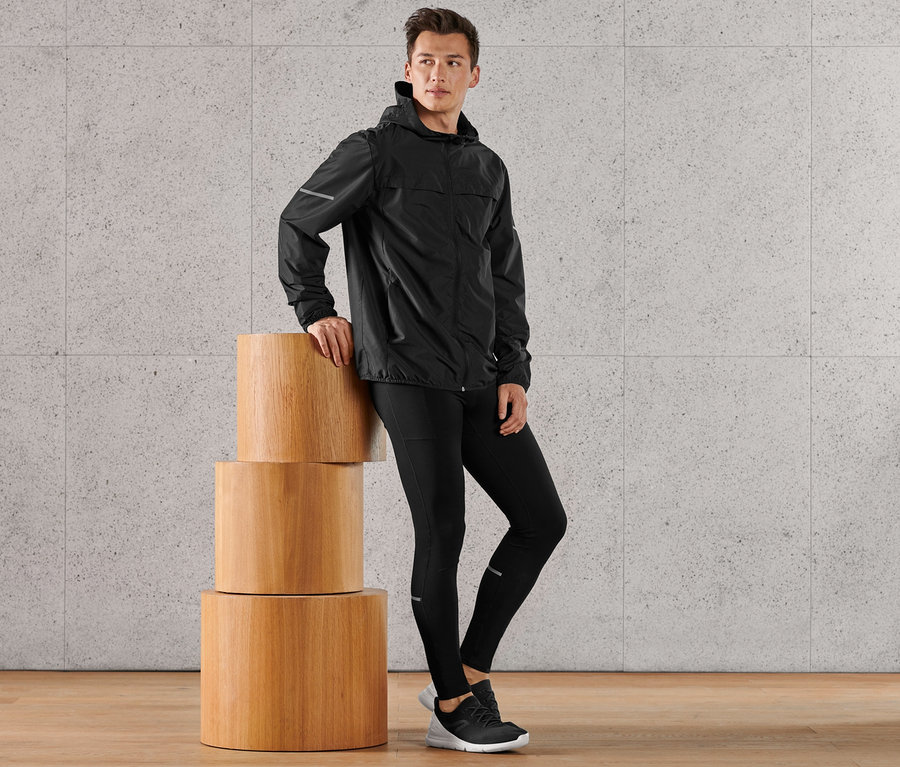 Mann in schwarzer Laufjacke und schwarzer Performance-Lauftight, schwarz, lehnt an drei Holzzylindern.