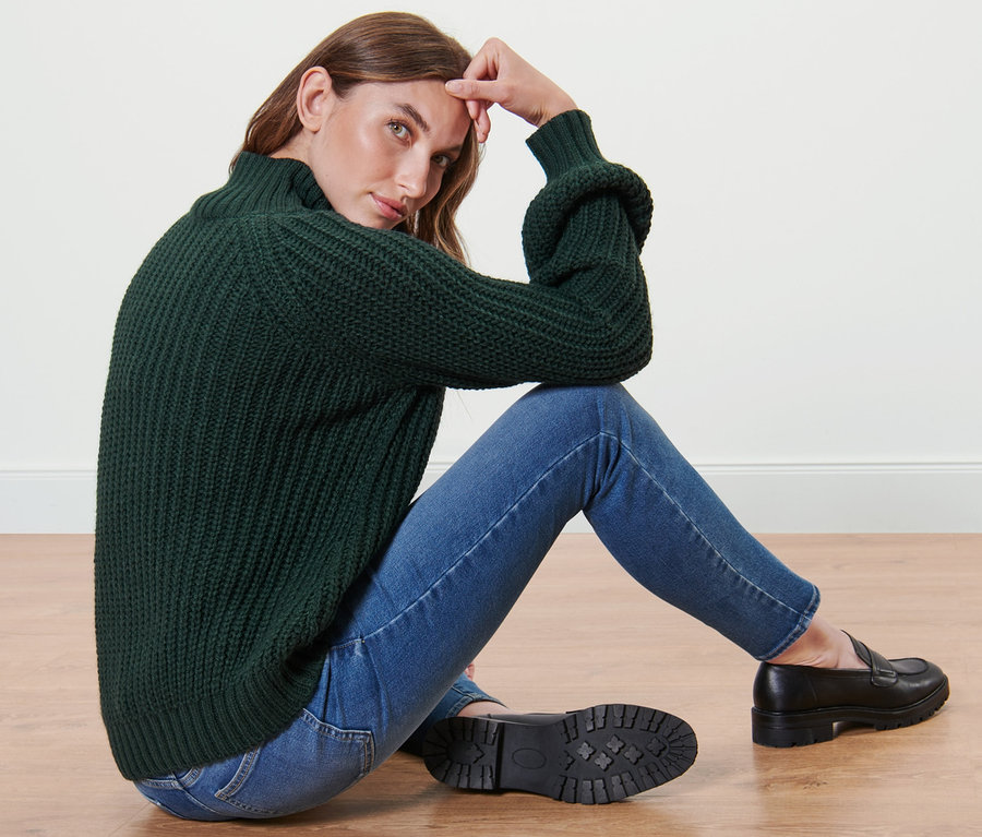 Frau sitzt auf dem Boden und trägt einen dunkelgrünen Strickpullover mit Stehkragen, Jeans und schwarze Slipper.