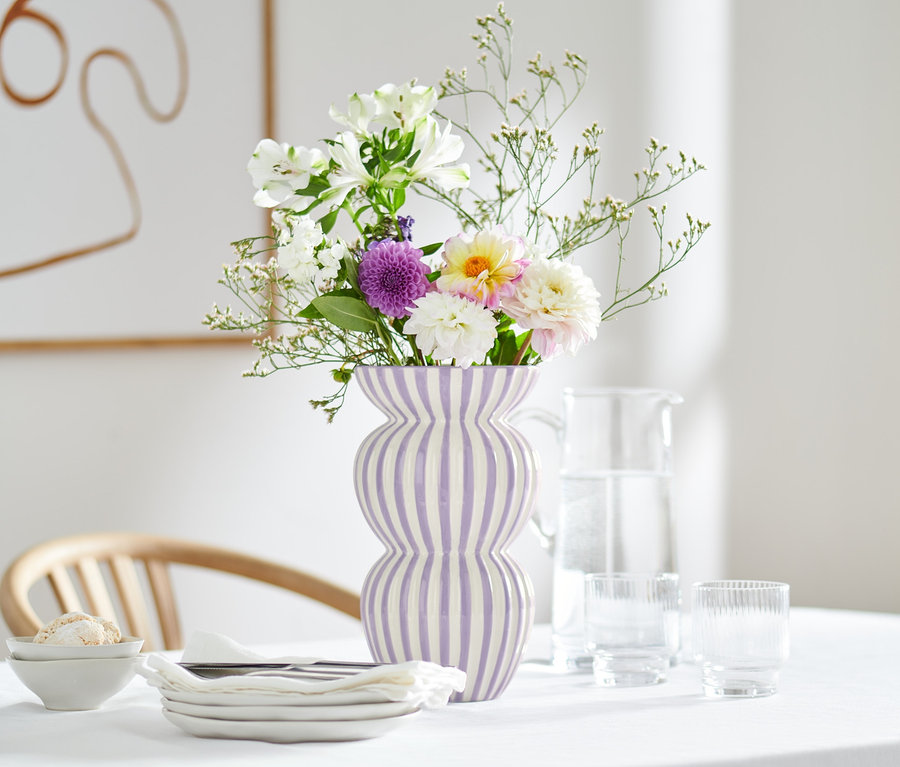 Blumen in großer Vase mit lila Streifen auf Tisch mit Blumendecke.