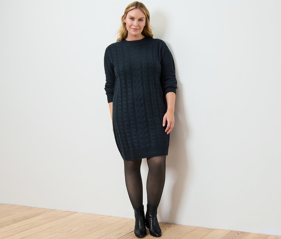 Frau posiert in Strickkleid mit Zopfmuster und 50-DEN-Feinstrumpfhose in transparenter Optik.