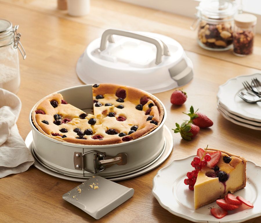 Ein angeschnittener Kuchen mit Früchten in einer Springform mit Transportdeckel neben einem Teller mit Kuchenstück.