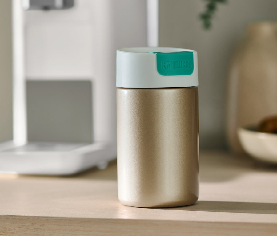 Goldener Kambukka To-Go-Becher steht auf einem Tisch neben einer Kaffeemaschine.
