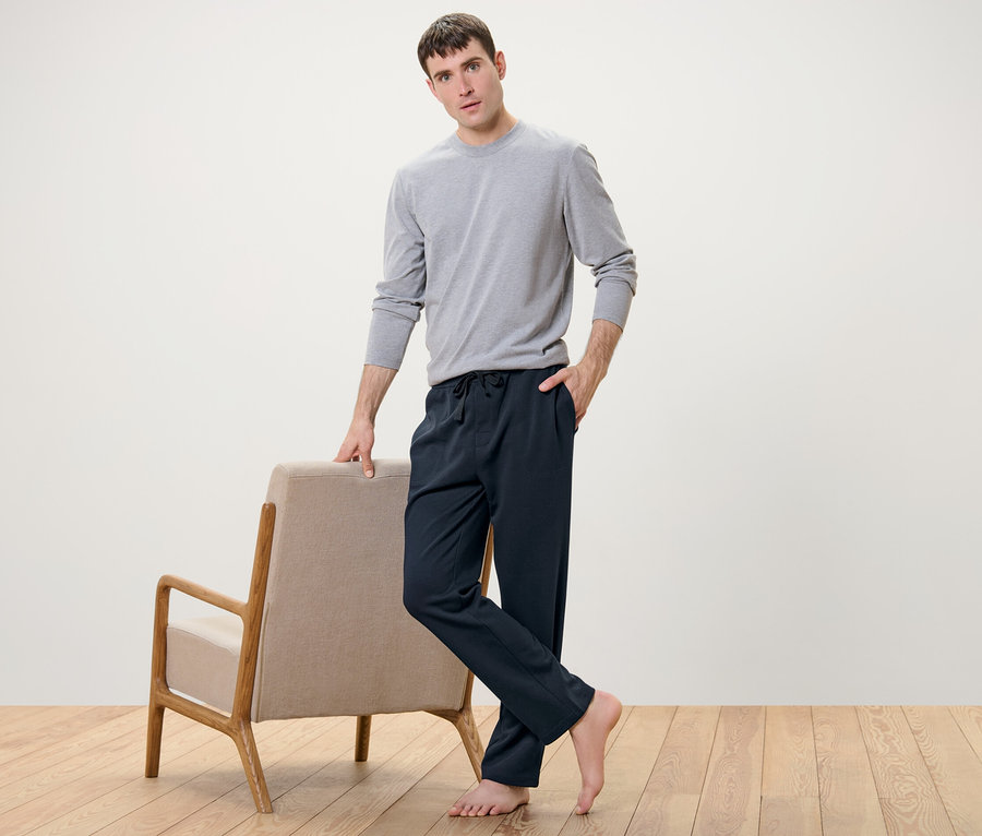 Mann posiert neben Sessel und trägt Homewearhose und graues Shirt.