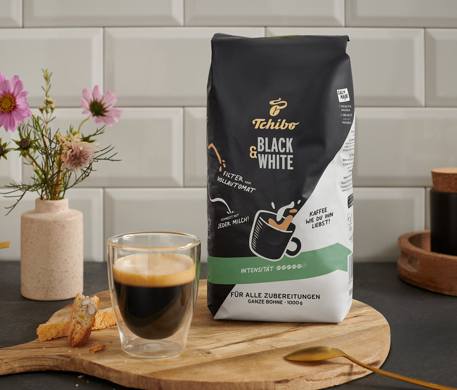 Ein Glas Kaffee und Gebäck stehen neben einer Packung BLACK & WHITE - 1 kg Ganze Bohne auf einem Holzbrett.