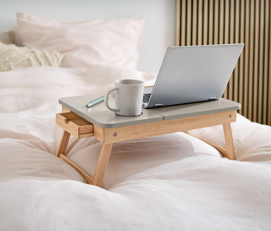 Auf einem Bett steht ein Laptoptisch aus Holz, auf dem sich ein Laptop, eine Tasse und ein Stift befinden.