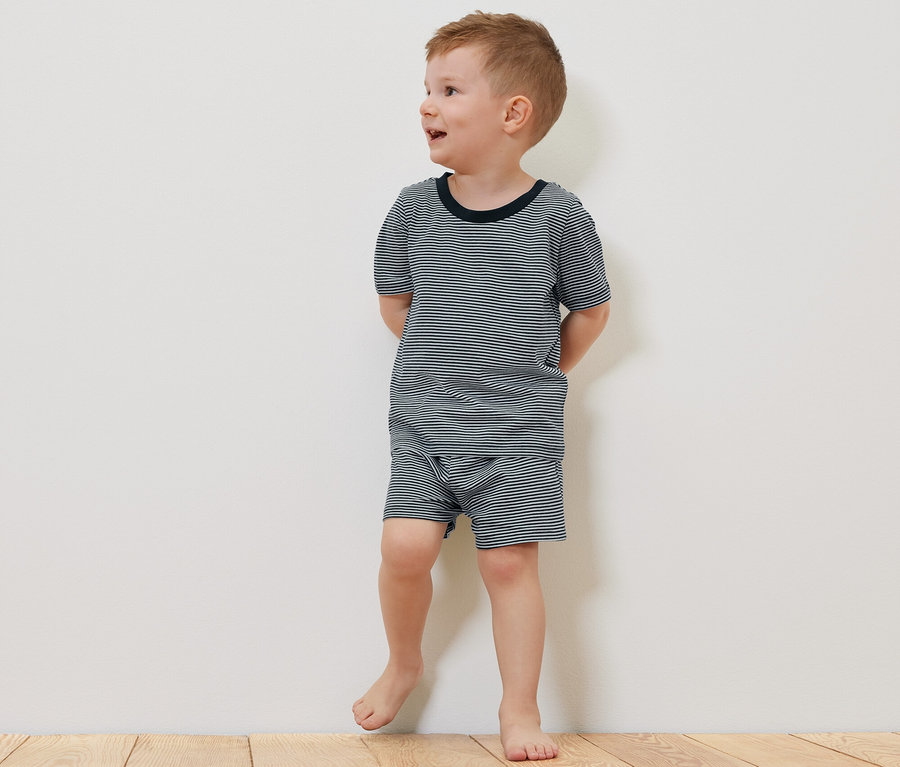 Ein Junge in einem kurzärmeligen 2 Kinder-Pyjama, kurz, steht barfuß auf einem Holzboden neben einem Teddybär.