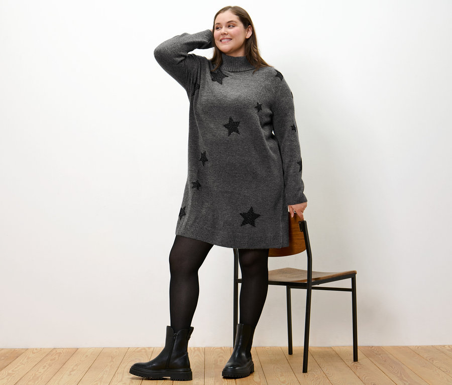 Frau posiert in einem Strickkleid mit Rollkragen und schwarzen Sternen, dazu trägt sie schwarze Stiefel und Strumpfhosen.