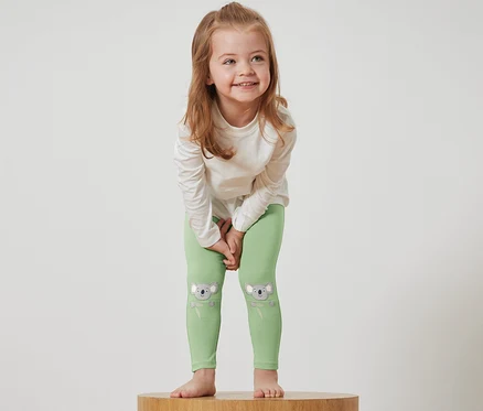 Mädchen steht auf einem Holzsockel und trägt grüne Leggings mit Koalabildern und ein weißes T-Shirt.