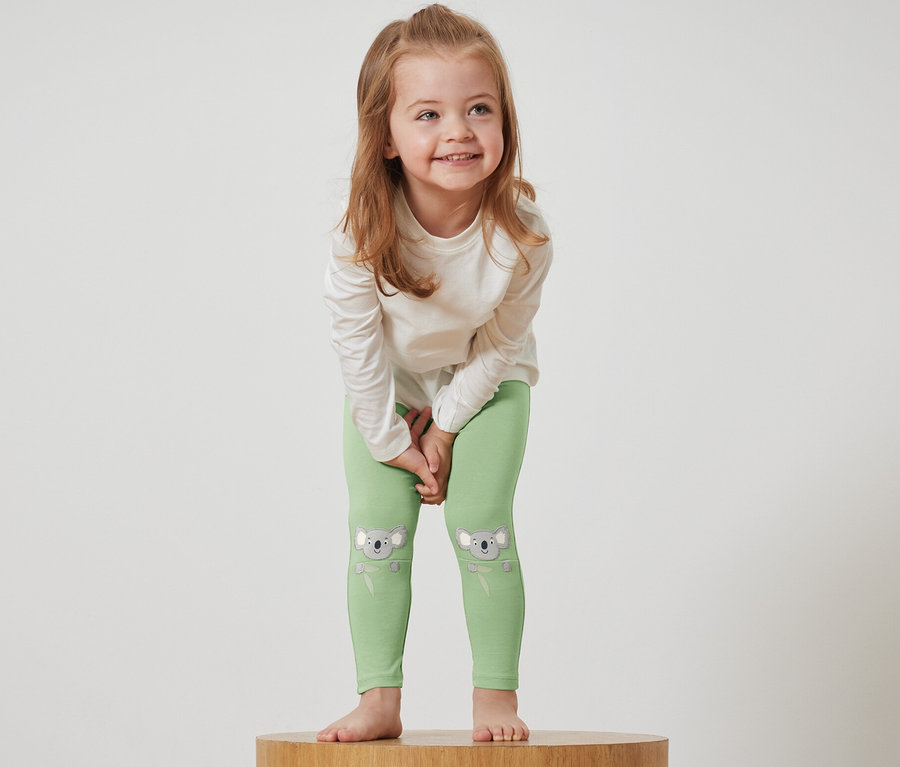 Mädchen steht auf einem Holzsockel und trägt grüne Leggings mit Koalabildern und ein weißes T-Shirt.
