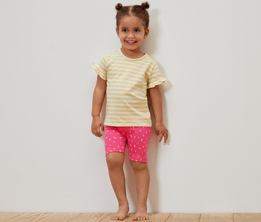 Ein lächelndes Mädchen steht an einer Wand und trägt ein gelb gestreiftes T-Shirt und eine rosa Radlerhose mit Muster. Das Mädchen trägt 2 Kinder-Radler-Leggings, pink und gelb.