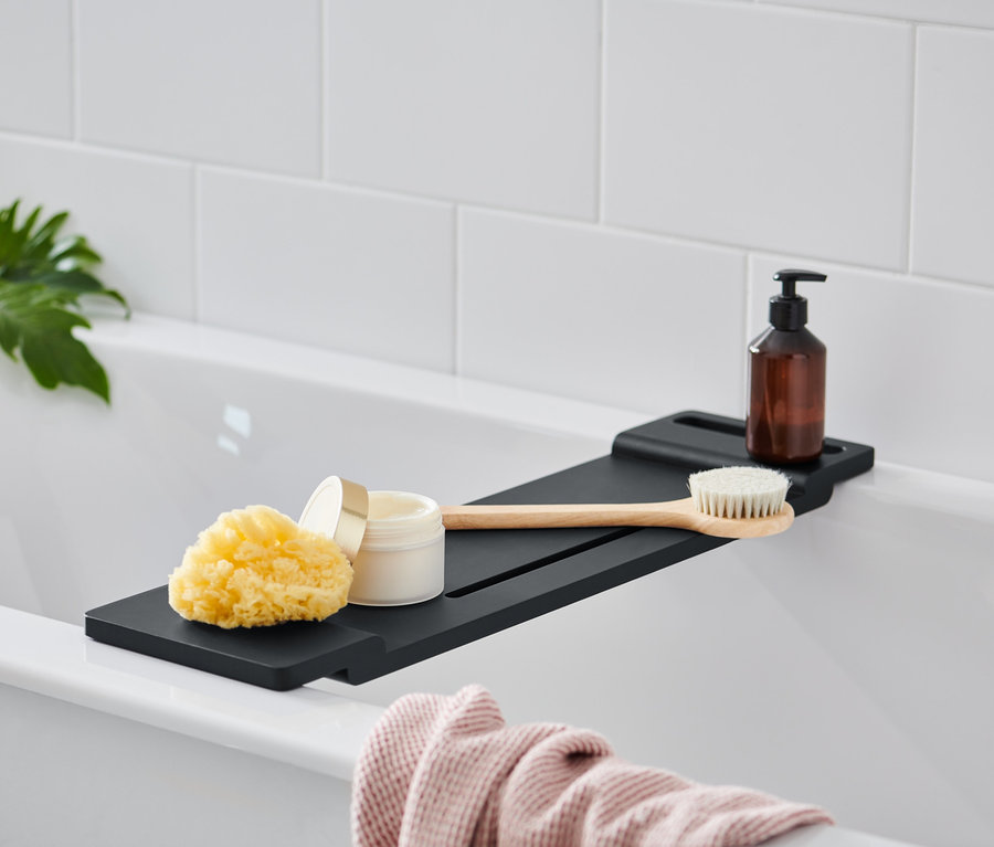 Auf einer Badewanne befindet sich ein schwarzes Badewannen-Tablett, auf dem ein Schwamm, ein Tiegel mit Creme, eine Bürste und eine braune Flasche mit Pumpspender liegen.