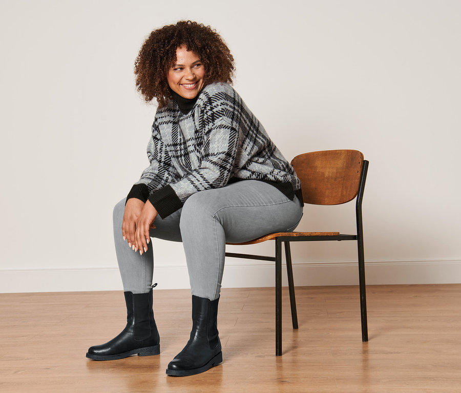 Frau sitzt auf einem Stuhl und trägt einen Strickpullover mit Rollkragen, hellgraue Treggings und Lederboots.