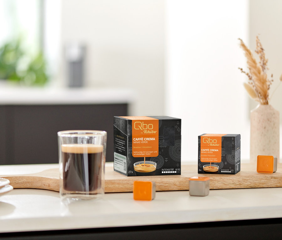 Ein Glas Kaffee mit Crema, zwei Qbo Caffè MINAS VERDE Packungen (eine für 27 Kapseln, eine für 8 Kapseln) und zwei lose Qbo Kapseln (eine orange, eine grau) sind auf einem Holzbrett auf einer Küchenzeile arrangiert. Eine Vase mit Trockengras und eine Schale mit Keksen sind im Hintergrund.