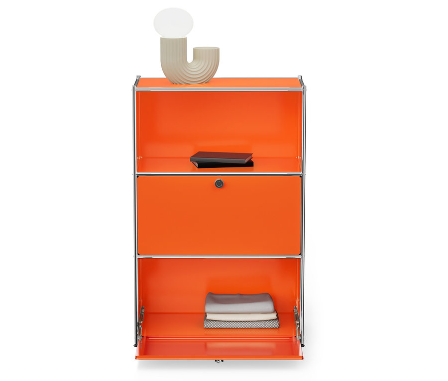 Orangefarbener Schrank mit drei Fächern und silbernem Rahmen. Auf dem oberen Fach befinden sich eine hellbeige Lampe und ein schwarzes Buch. Das untere Fach ist geöffnet und enthält gefaltete Stoffe.