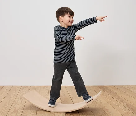 Ein Junge steht auf einem hölzernen Balance Board, lacht und zeigt mit dem Finger.