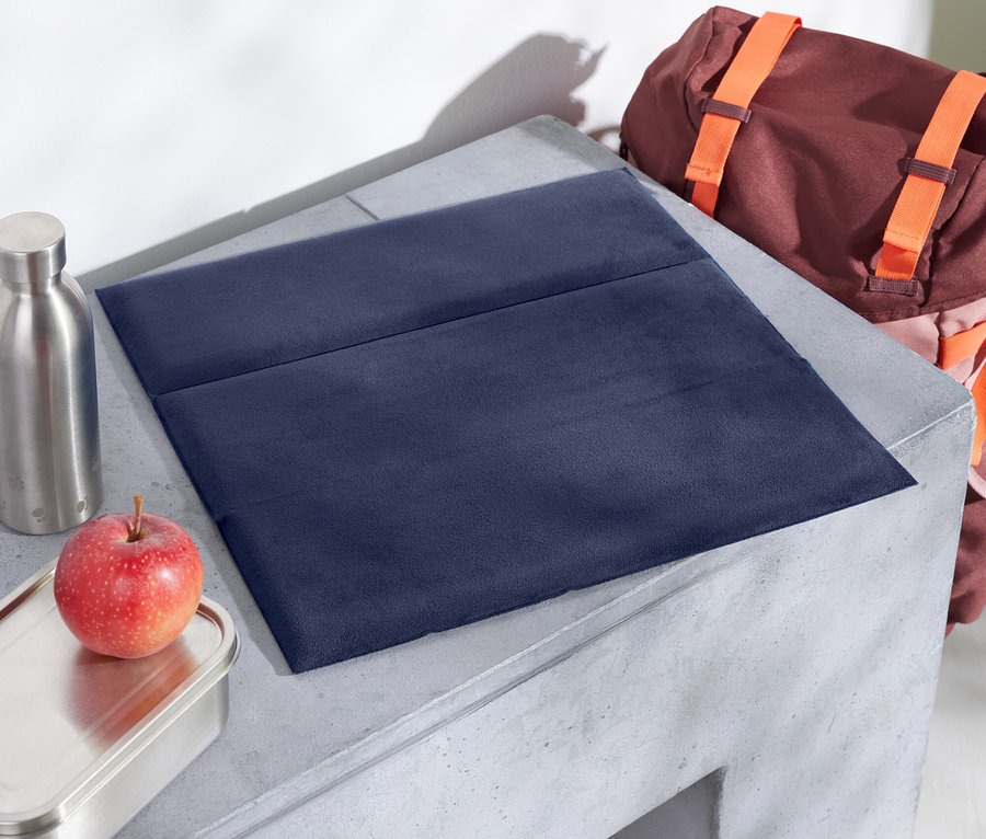 Auf einem Betontisch liegen eine blaue Stoffunterlage, ein Apfel, eine Edelstahldose und eine Flasche. Im Hintergrund befindet sich ein brauner Rucksack mit orangefarbenen Trägern.