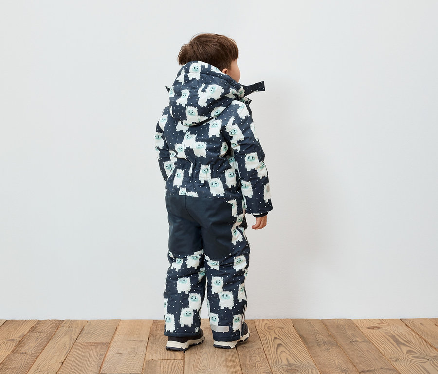 Junge steht mit dem Rücken zur Kamera und trägt einen blau-weißen Overall mit Kapuze und Schneemannmuster.