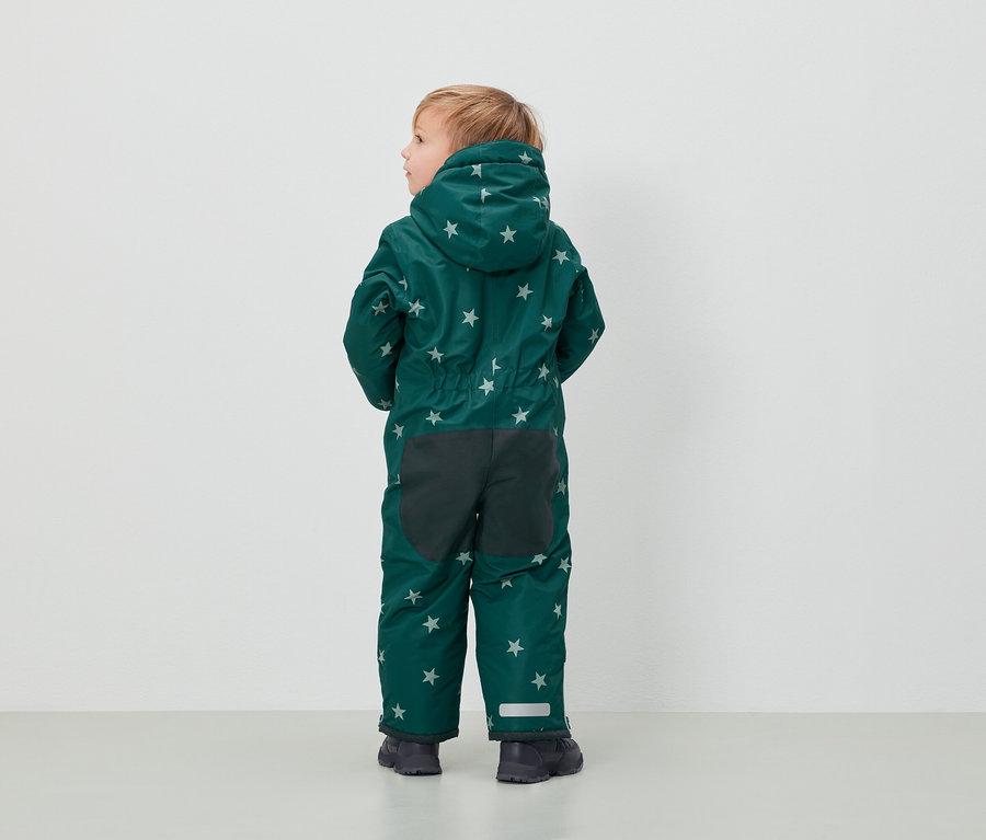 Ein Junge in einem grünen Overall mit Sternmuster steht mit dem Rücken zur Kamera.