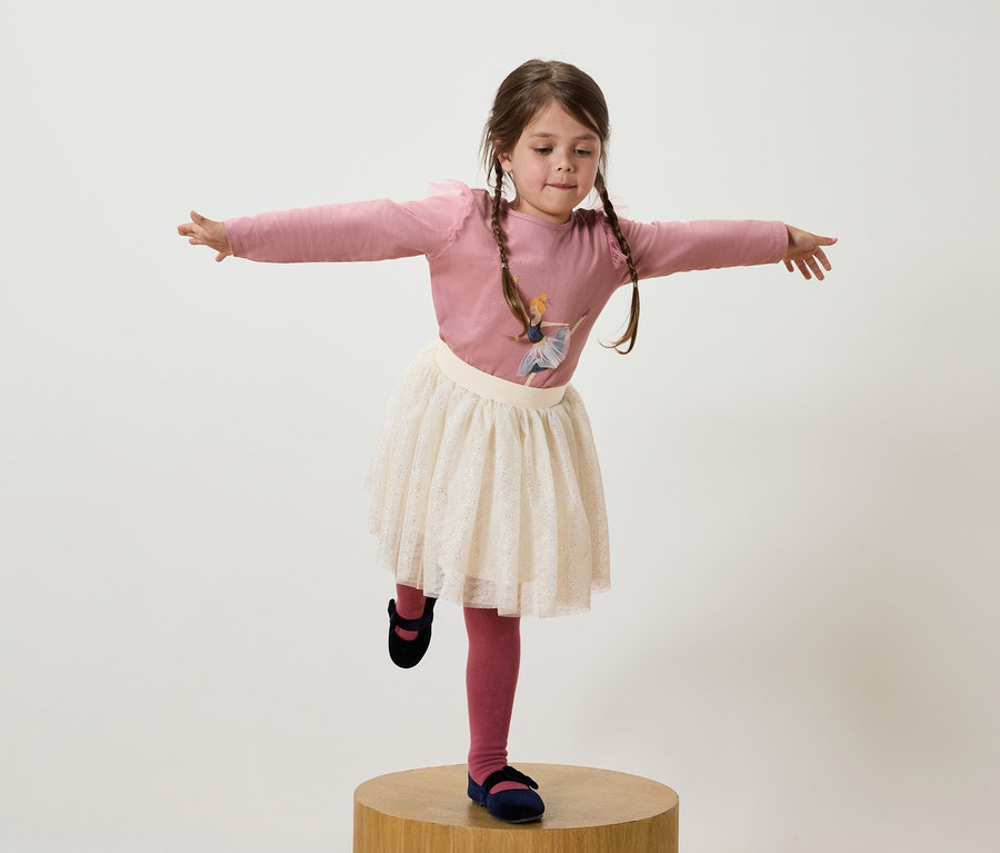 Mädchen mit braunen Zöpfen steht auf einem Bein auf einem Holzsockel, trägt ein rosa T-Shirt, einen weißen Rock und dunkelblaue Schuhe.