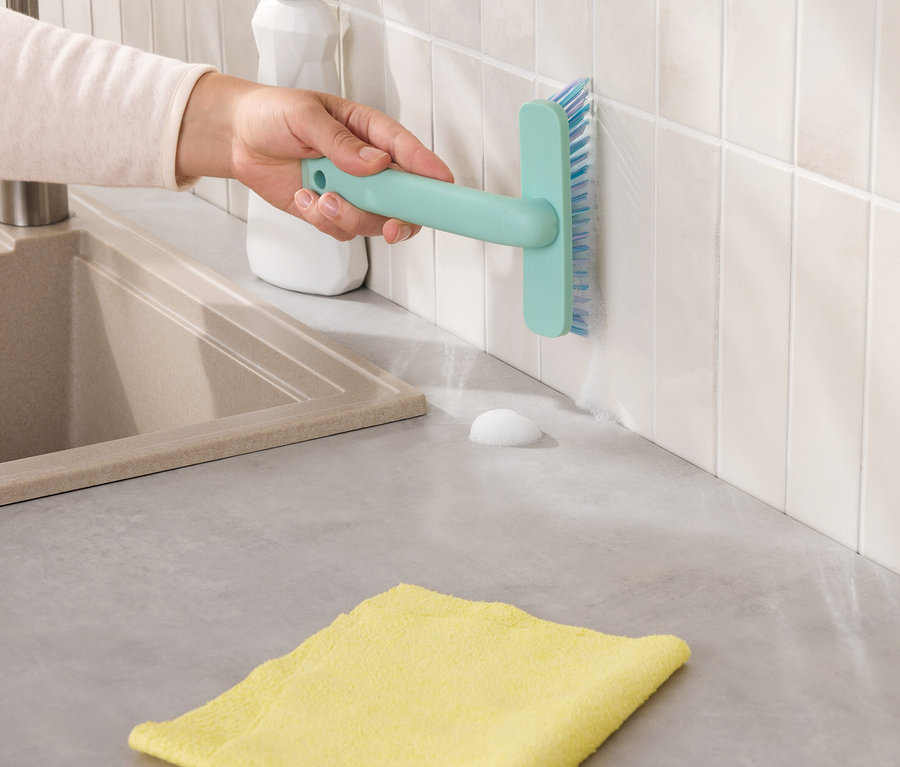 Eine Hand reinigt weiße Fliesen mit einer hellblauen Bürste, neben einem beigen Waschbecken und einem gelben Tuch.