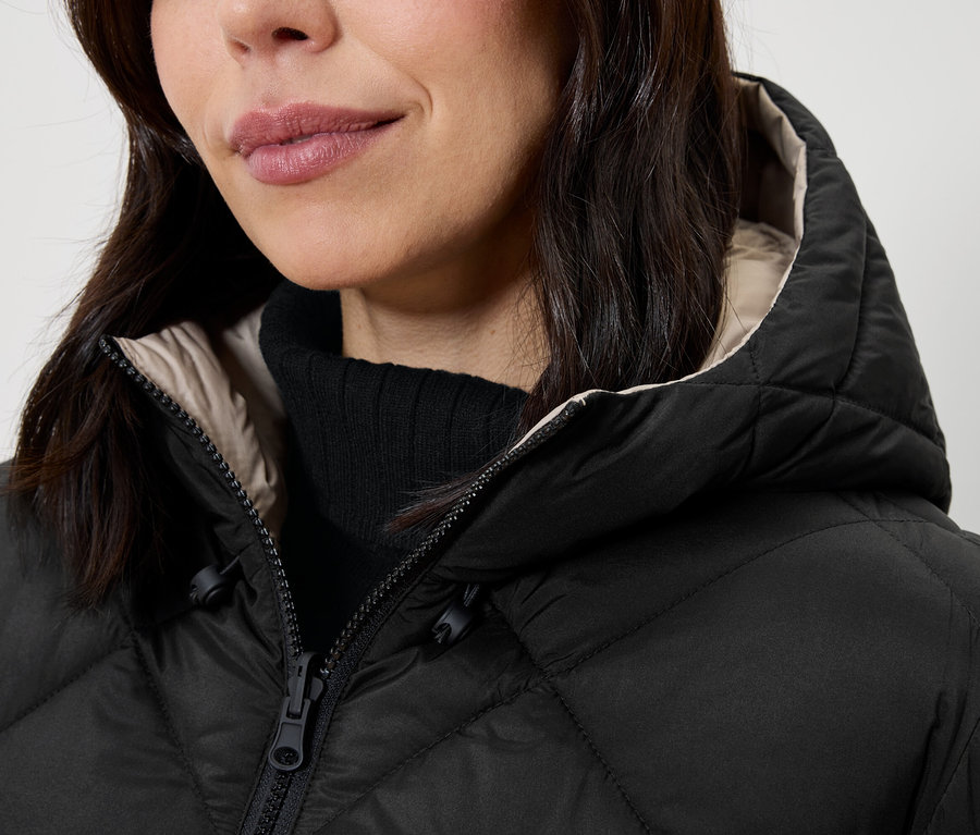 Detailaufnahme einer Frau mit einer schwarzen Steppjacke mit Kapuze und einem schwarzen Rollkragenpullover.