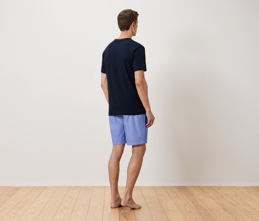 Ein Mann steht mit dem Rücken zur Kamera, er trägt ein marineblaues T-Shirt und hellblaue Shorts.