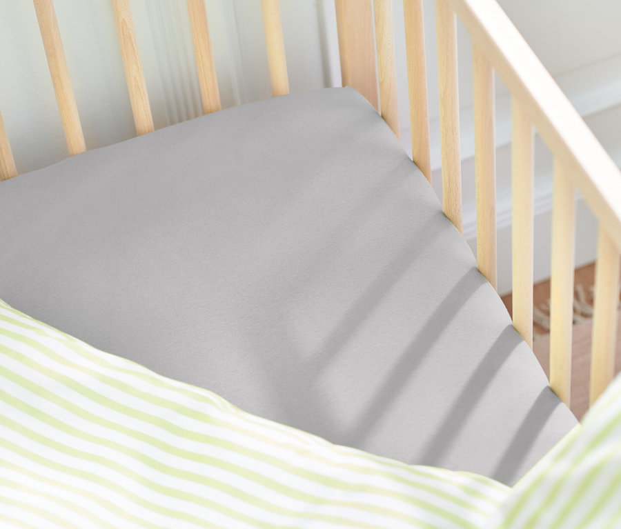 Detailaufnahme eines Kinderbetts mit grauem Spannbettlaken und einem Kissen mit grünen Streifen.