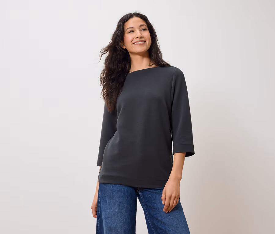 Frau mit dunklem Haar posiert in einem grauen Sweatshirt mit Dreiviertelärmeln und blauen Jeans.