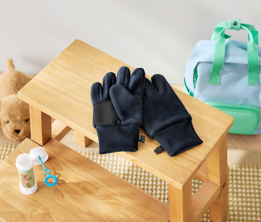 Auf einem Holzschemel liegen dunkelblaue Handschuhe, daneben eine Seifenblasenflasche und ein Plüschhund. Im Hintergrund befindet sich ein hellblauer Rucksack mit mintgrünen Details.