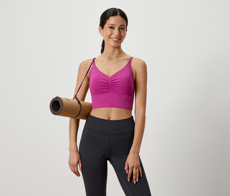 Frau mit brauner Yogamatte über der Schulter, gekleidet in einem pinkfarbenen Sportoberteil und schwarzen Leggings.