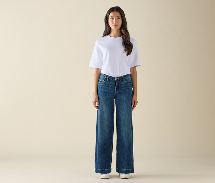 Frau steht in weißem T-Shirt, blauen Schlaghosen und weißen Turnschuhen.