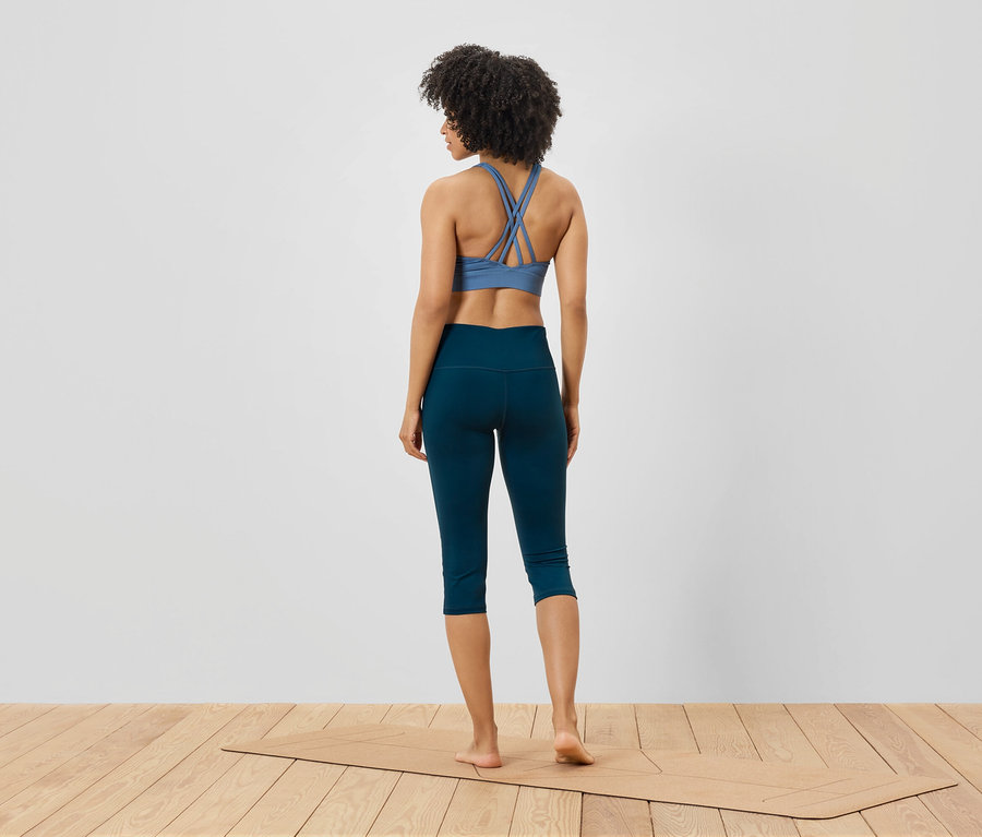 Frau steht mit dem Rücken zur Kamera, trägt einen blauen Sport-BH und marineblaue Leggings und steht auf einer Yogamatte.