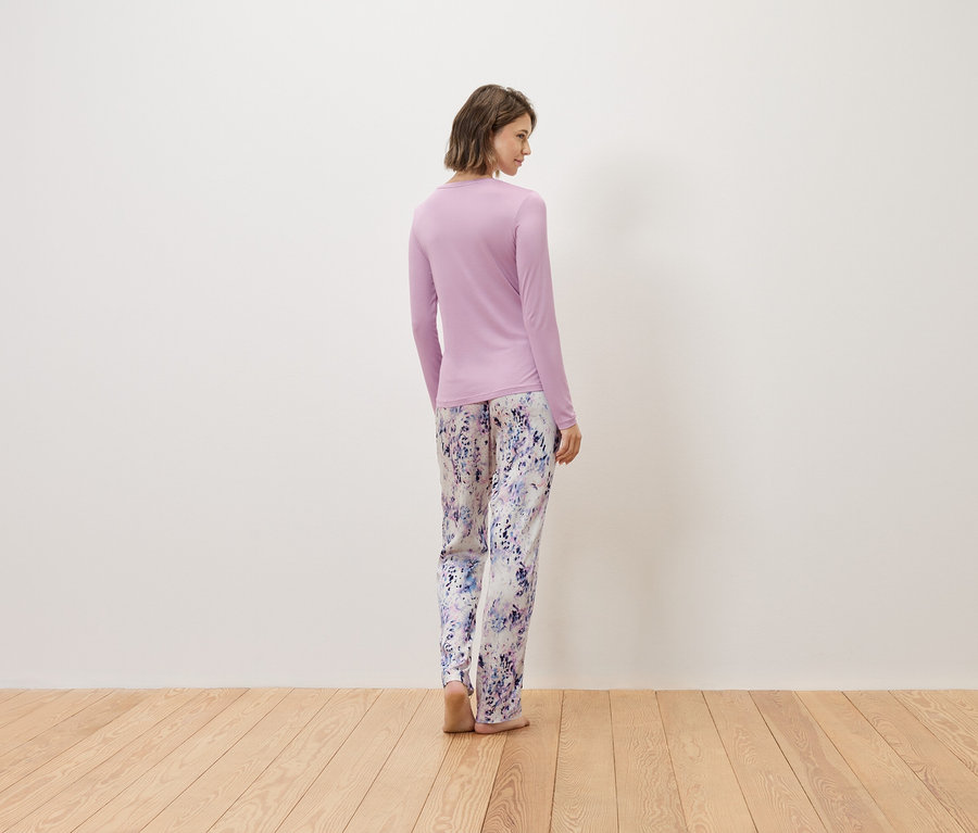 Eine Frau steht mit dem Rücken zur Kamera, sie trägt einen langärmligen lila Pullover und eine lange Pyjamahose mit Blumenmuster.