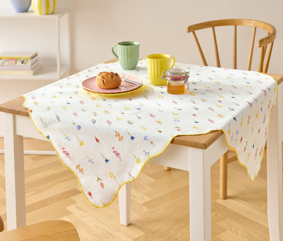 Ein Tisch mit einer weißen Tischdecke mit Blumenmuster, zwei Tassen, einem Teller mit einem Brötchen und einem Glas Honig.
