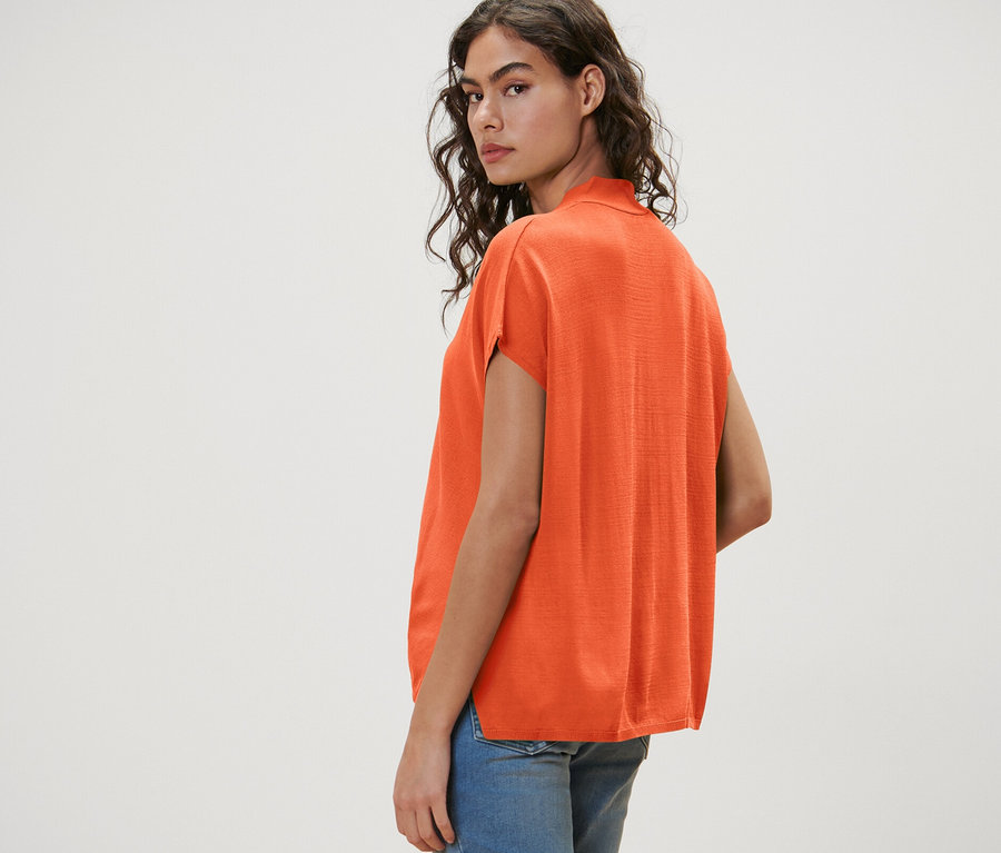 Frau blickt über ihre Schulter und trägt einen orangefarbenen, kurzärmligen Cardigan und Jeansshorts.