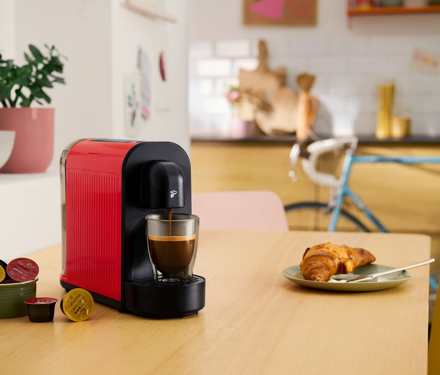 Eine rote Kaffeekapselmaschine füllt eine Tasse mit Kaffee. Auf dem Tisch liegen ein Croissant, eine Schale mit Kaffeekapseln und eine Topfpflanze.