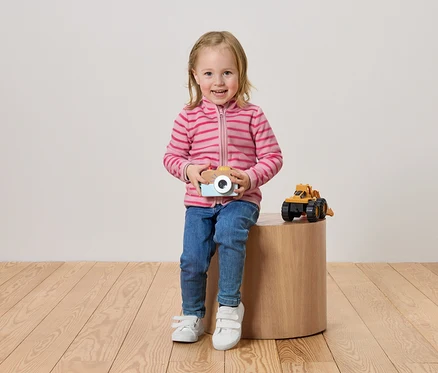 Ein kleines Mädchen sitzt auf einem Holzhocker und hält eine Spielzeugkamera in den Händen. Neben ihr steht ein gelber Spielzeug-Bagger.