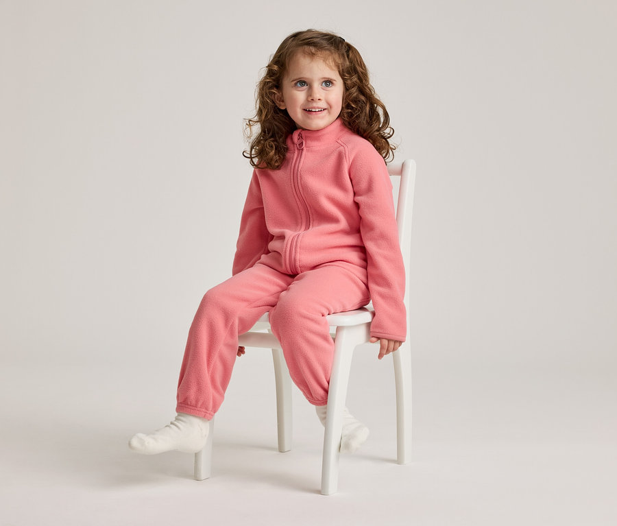 Ein Mädchen mit lockigem Haar sitzt auf einem weißen Stuhl und trägt einen rosa Fleece-Overall und weiße Socken.
