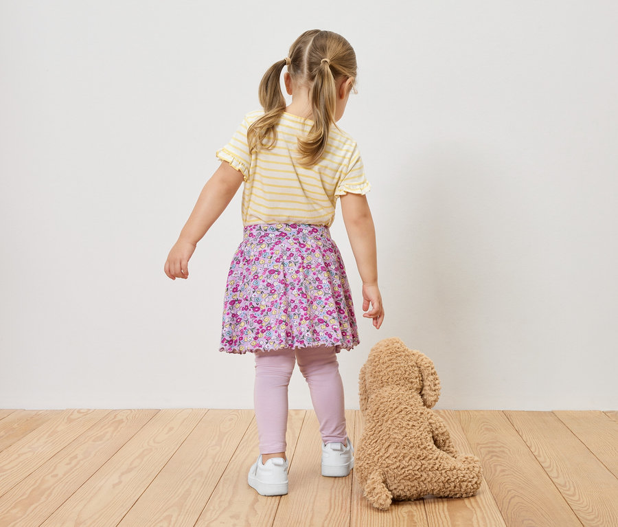 Blick von hinten auf ein Mädchen mit Zöpfen in einem gelb-weiß gestreiften Oberteil, einem geblümten Rock, Leggings und Turnschuhen, neben einem Teddybär.