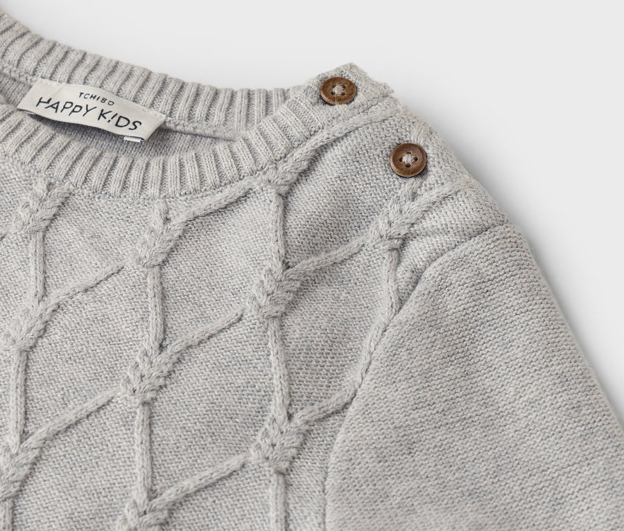 Detailansicht eines hellgrauen Strickpullovers mit Zopfmuster und zwei Knöpfen an der Schulter.