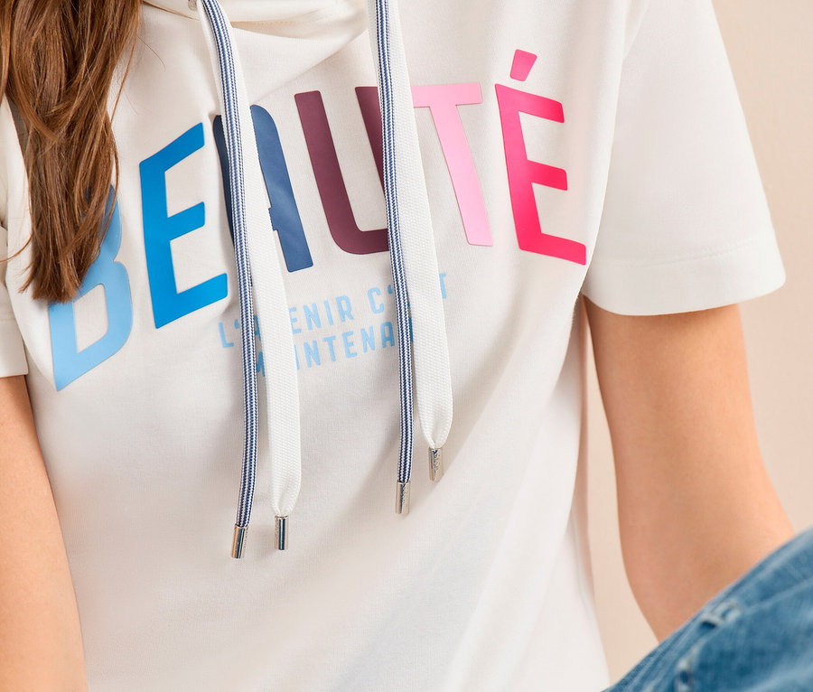 Sitzende Frau in weißem CECIL Hoodie-Shirt mit kurzen Ärmeln, farbigem BEAUTÉ-Aufdruck und blauen Jeans.