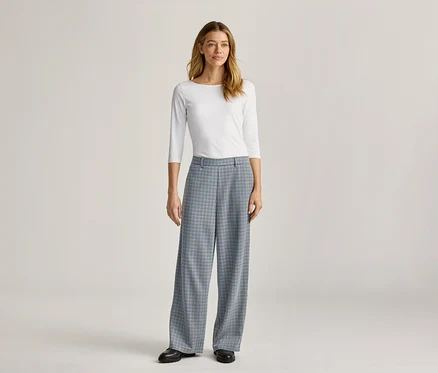 Eine Frau steht in einem weißen, langärmeligen Oberteil und einer weiten, blau karierten Hose mit schwarzen Schuhen.