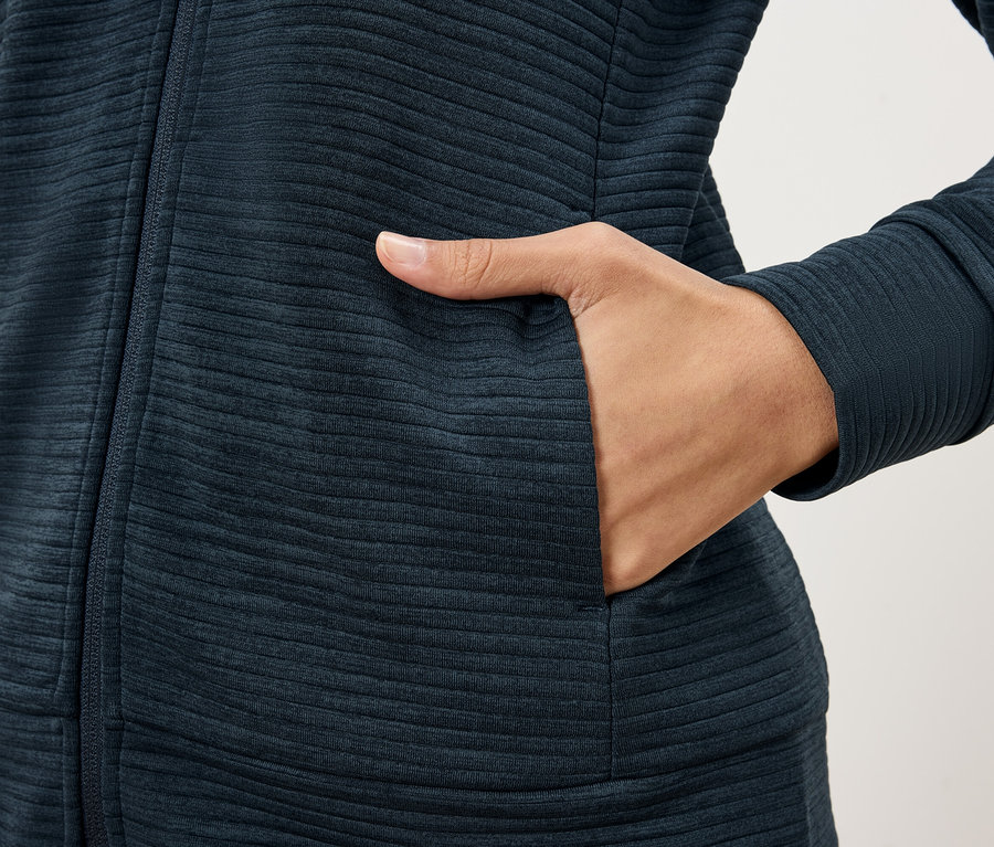 Nahaufnahme einer Person, die eine dunkelblaue strukturierte Jacke trägt und ihre Hand in eine Jackentasche steckt.