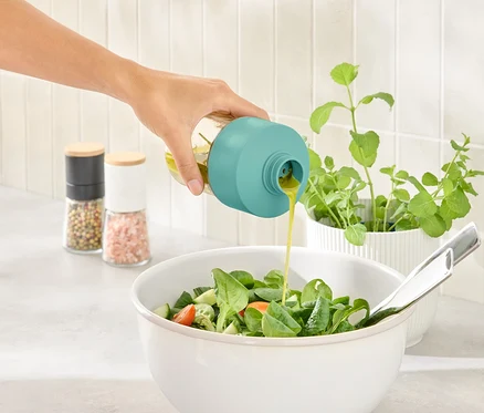 Eine Hand gießt Dressing aus einer Flasche mit türkisfarbenem Verschluss in eine Salatschüssel.
