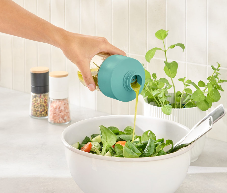 Eine Hand gießt Dressing aus einer Flasche mit türkisfarbenem Verschluss in eine Salatschüssel.