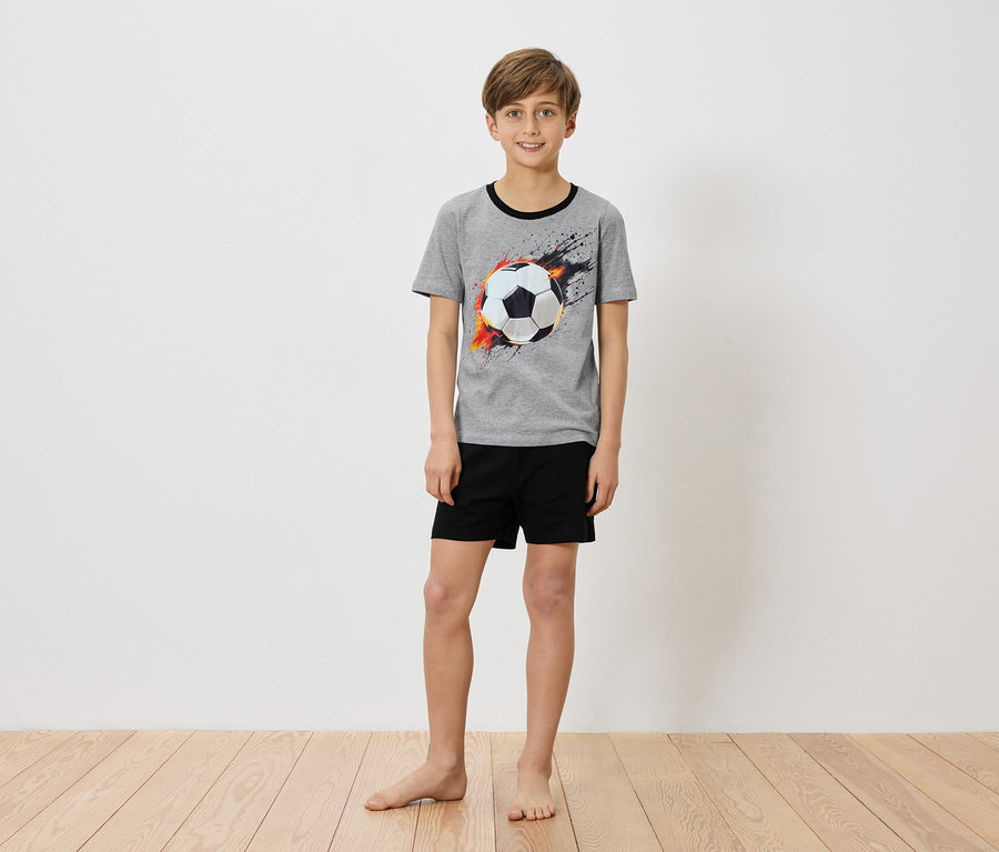 Ein Junge steht barfuß vor einer weißen Wand und trägt ein graues T-Shirt mit einem Fußballaufdruck und schwarzen Shorts.